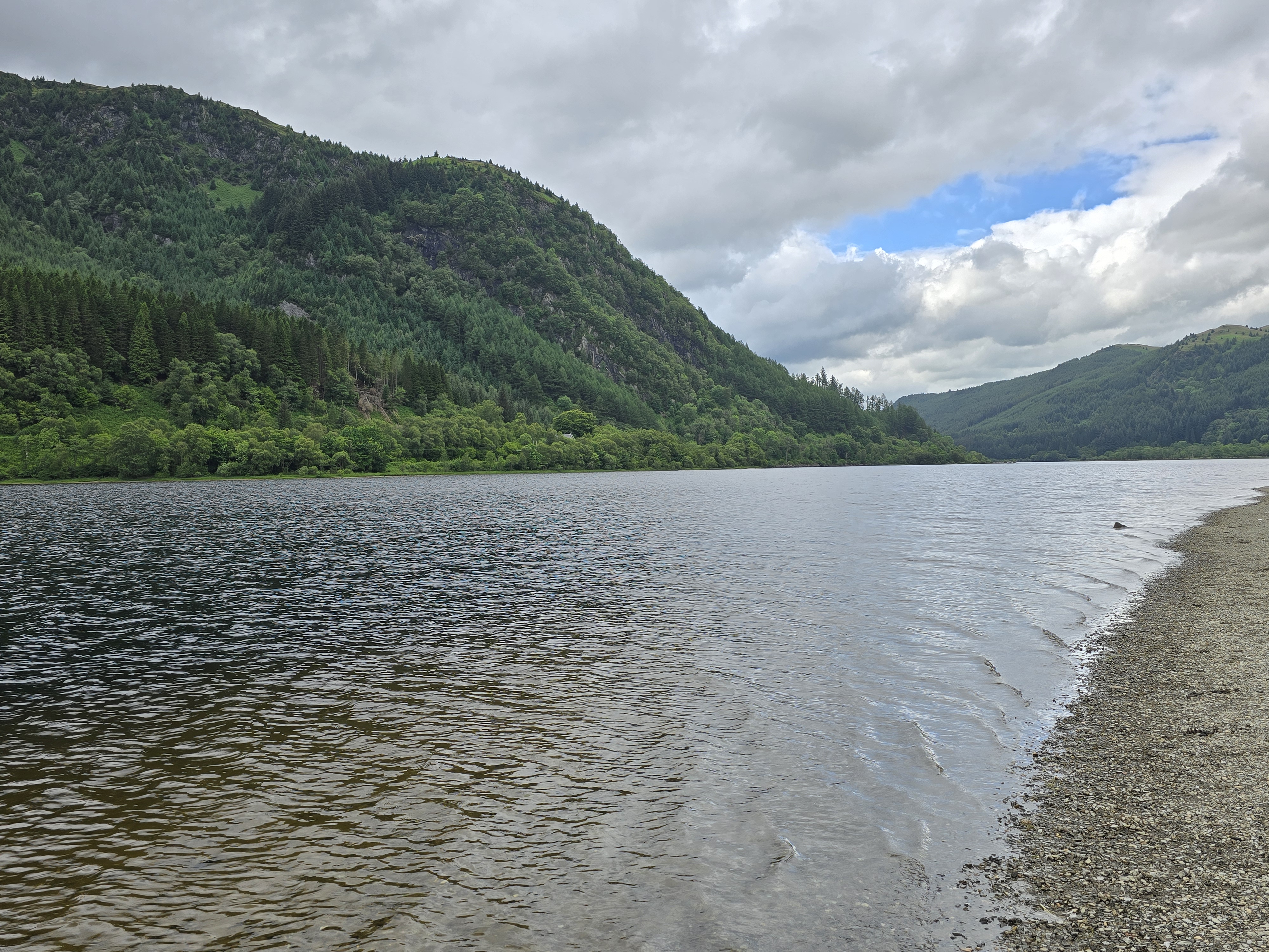 Still dark water of a Scottish loch with green forested hills rolling down to the shore under a partly cloudy sky