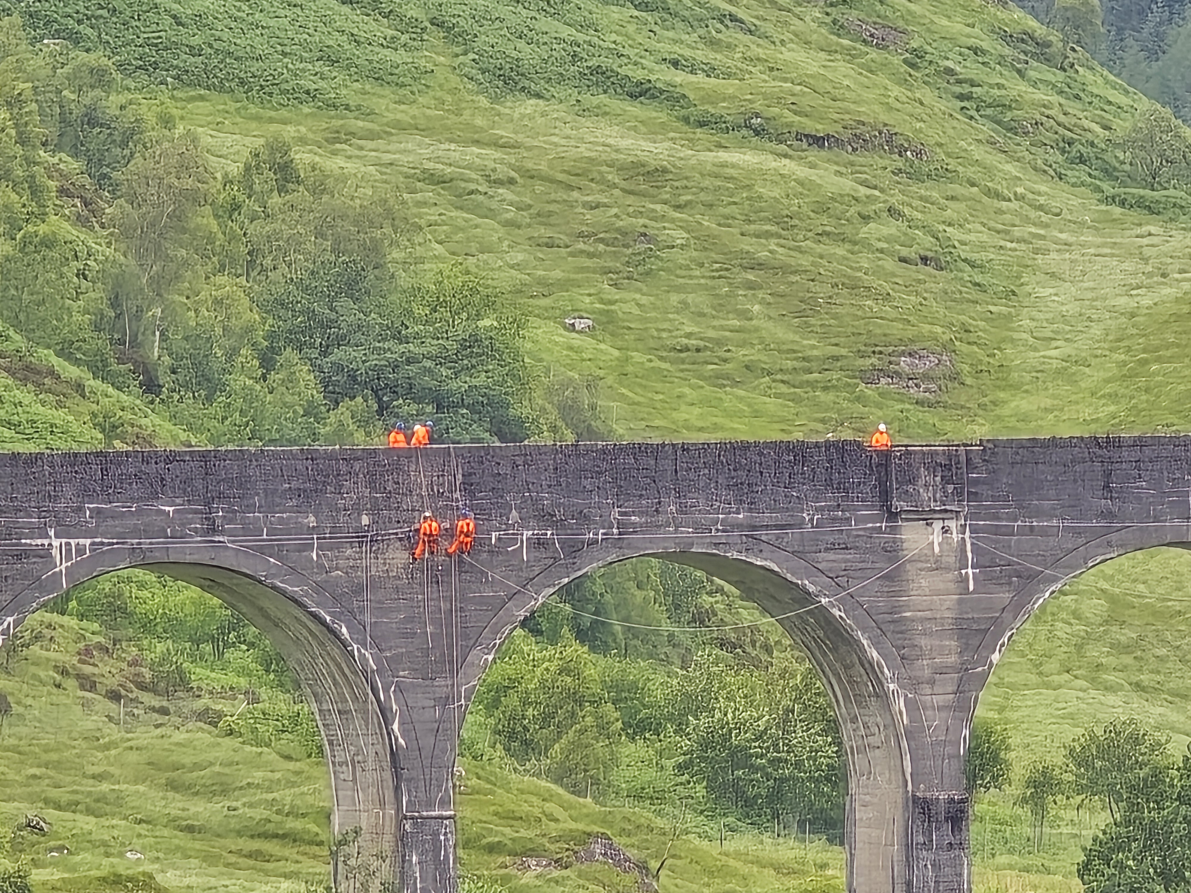 Glenfinnan Viaduct with maintenance workers in orange high-visibility vests working on the stone arches, surrounded by lush green Scottish hills