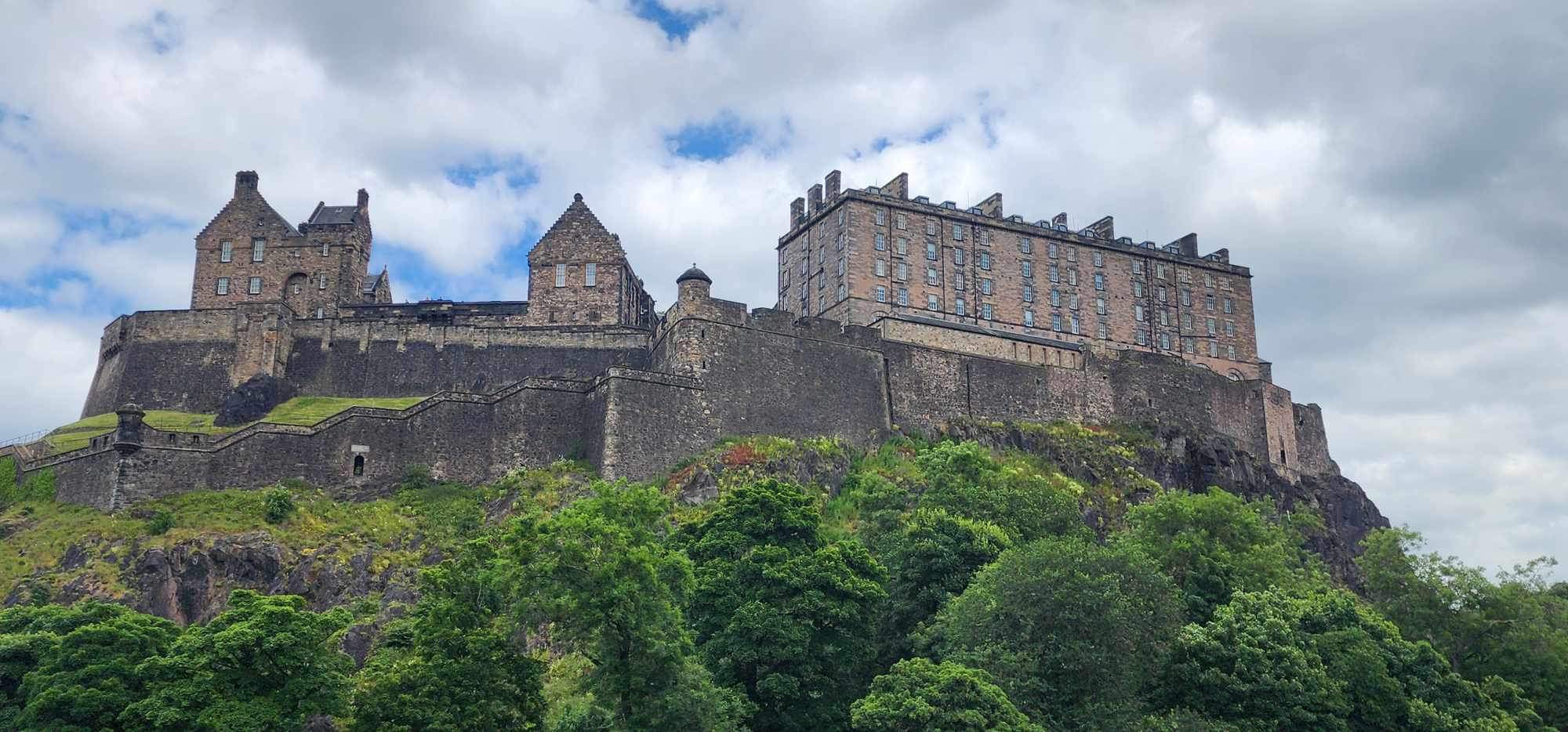 Edinburgh Castle viewed dramatically from below, perched on volcanic rock with green trees in the foreground