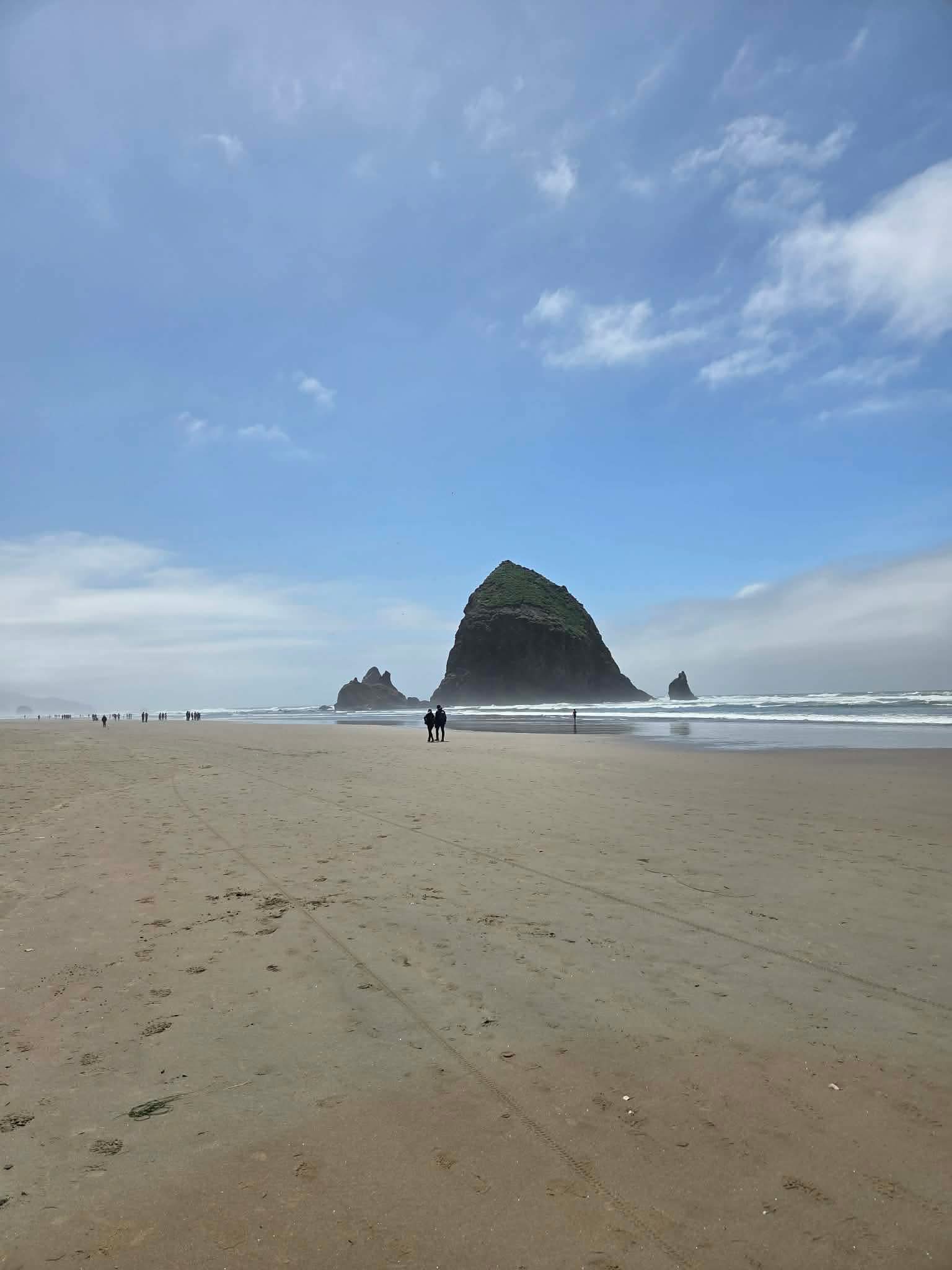 Haystack Rock rising dramatically from the wide flat sand of Cannon Beach Oregon under a partly cloudy sky with small figures walking in the distance